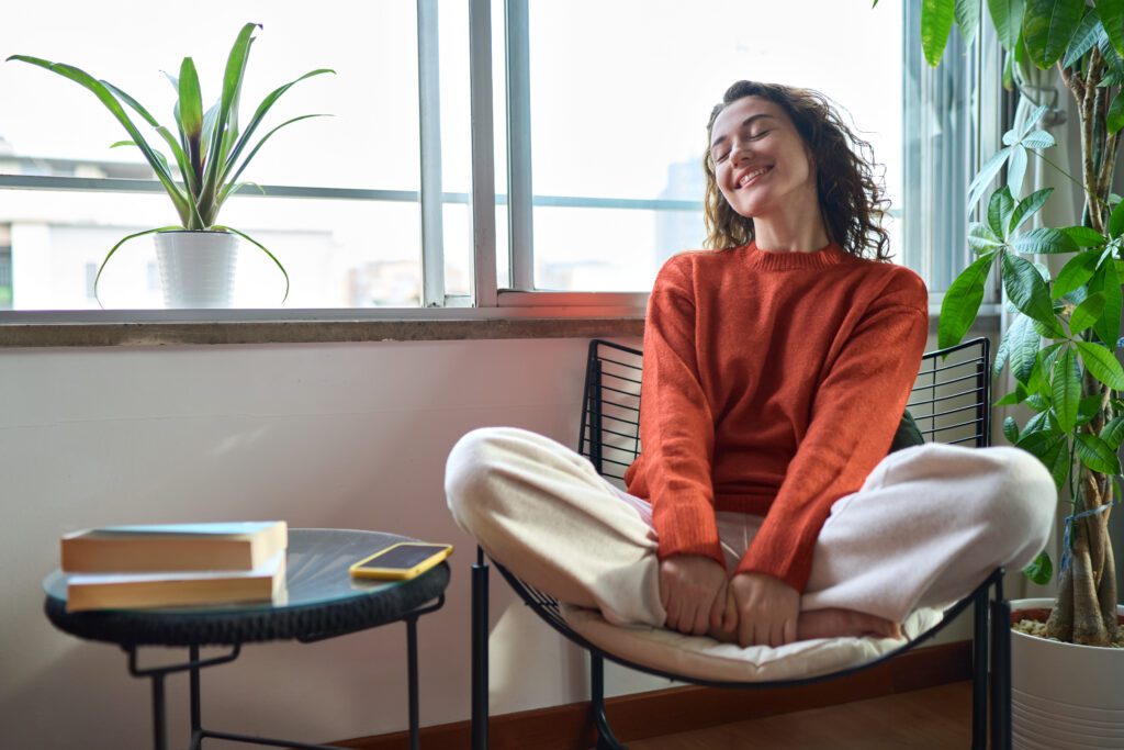 A young woman smiling confidently in a chair beside a window.